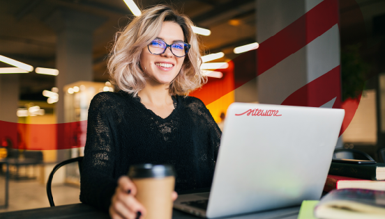 Jovem mulher sorridente com cabelo cacheado e cabelos loiros claros trabalhando com laptop em cafeteria moderna, com iluminação aconchegante e decorada. Imagem usada como capa do Guia de Remuneração Variável da Siteware.