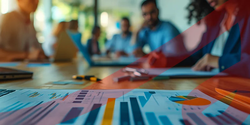 Reunião de negócios com pessoas ao fundo realizando análise de gráficos financeiros e estatísticos, ilustrando um conteúdo da Siteware sobre Programa de Participação nos Lucros e Resultados.