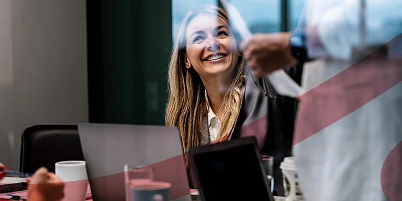 Mulher sorridente em reunião de negócios com laptop e café na mesa, ilustrando um conteúdo da Siteware sobre o que é bonificação salarial.