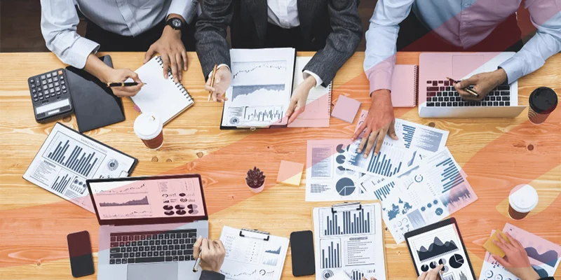 Vista de cima de equipe de negócios analisando gráficos e dados financeiros em laptops e documentos sobre mesa de madeira, ilustrando um artigo da Siteware sobre qual a diferença entre administração e gestão.