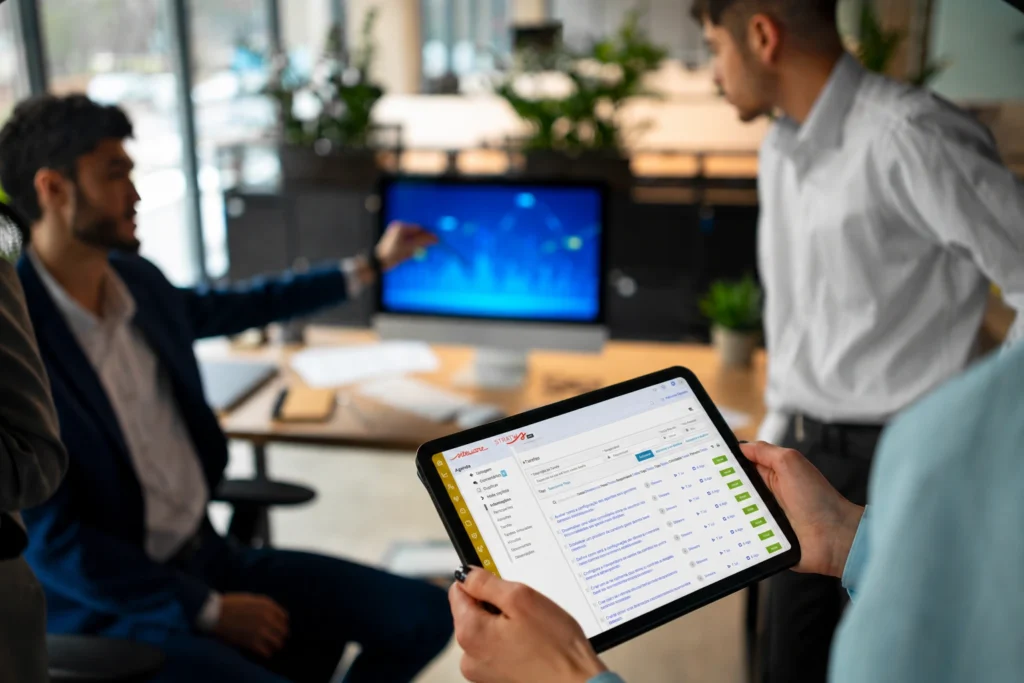 Profissional segurando tablet com a tela de gestão de tarefas do Stratws One, extraídas com o recurso de IA para reuniões do software. O fundo da imagem traz colaboradores discutindo indicadores exibidos em monitor.